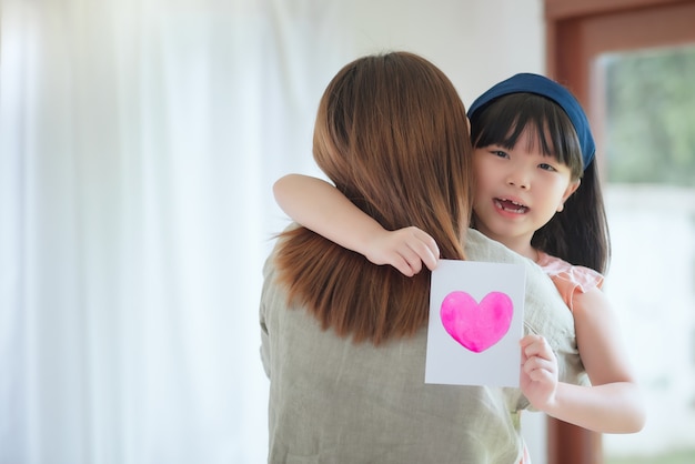 Asiatische Mutter umarmen ihre süße Tochter, die handgemachte Grußkarte mit buntem Herzsymbol geben, um sie zu Hause zu überraschen