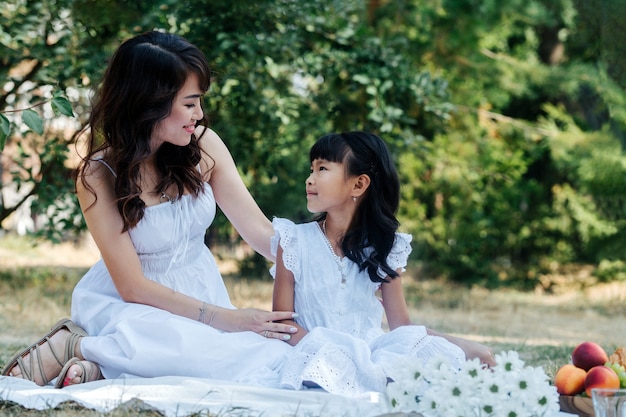 Asiatische Mutter und Tochter in weißen Kleidern beim Picknick in einem Park und genießen die letzten warmen Tage des frühen Herbstes in einem Baumschatten.