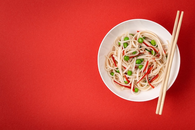 Asiatische Nudeln mit Gemüse, grünen Erbsen und rotem Pfeffer in weißer Schüssel mit Holzstäbchen, von oben. Asiatische vegetarische Nudeln auf rotem Hintergrund, Nahaufnahme, Draufsicht mit Platz für Text