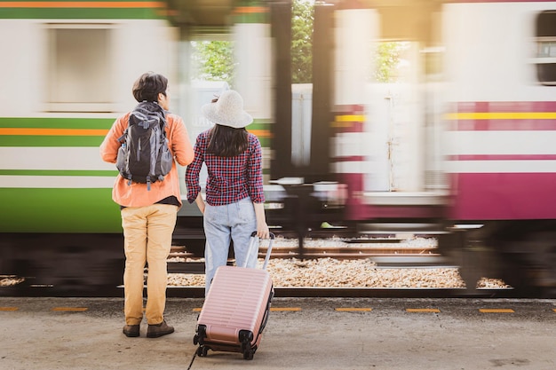 Asiatische Paartouristen mit Rucksäcken, die auf dem Bahnsteig im Bahnhof mit Zugbewegung stehen, sind Hintergrund.