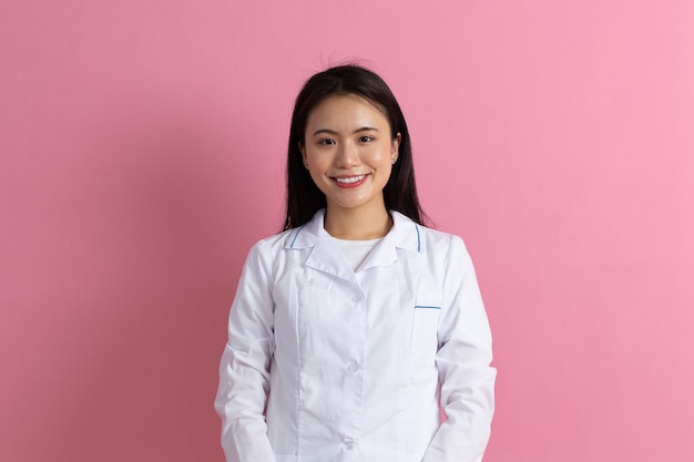 Asiatische positive Doktorfrau im weißen medizinischen weißen Kleid gegen rosa Hintergrundporträt