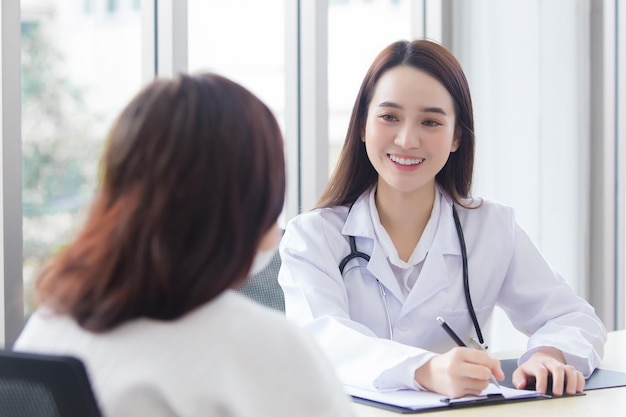 Asiatische professionelle Ärztin, die einen medizinischen Mantel trägt, spricht mit einer Patientin des Krankenhauses
