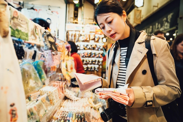 Foto asiatische reisefotografin, die souvenirs im japanischen taschentuchverkäufer auf dem brokatmarkt in kyoto japan kauft. chinesische touristin, die mehrere rosa mockets hält, die im geschenkeladen nishiki ichib auswählen