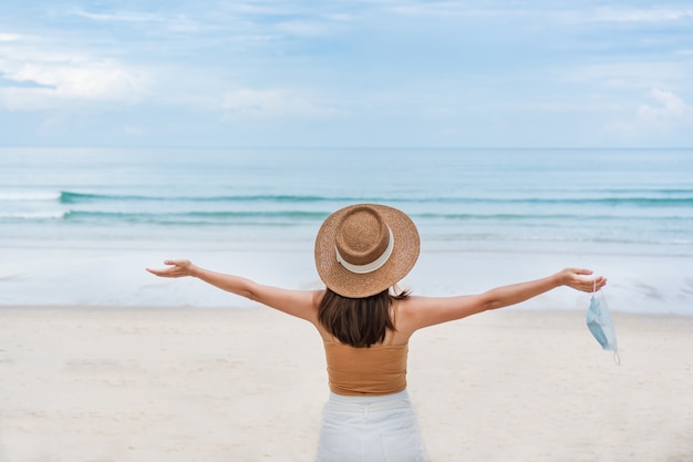 Asiatische reisende Frau nehmen medizinische Gesichtsmaske am Strand ab. Neues Normal-, Reise- und Urlaubskonzept