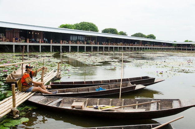 Asiatische Reisende thailändische Frau reisen besuchen und sitzen Bambusbrücke zum Fotografieren mit Holzboot, das mit Seerose im Teich im Garten des schwimmenden Marktes von Red Lotus in Nakhon Pathom Thailand schwimmt