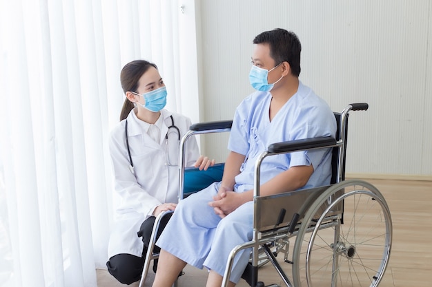 Asiatische Ärztin erklärt und schlägt einige Informationen mit einem männlichen Patienten vor, der auf einem Rollstuhl sitzt. Sie tragen eine medizinische Gesichtsmaske, während sie im Krankenhaus miteinander sprechen.