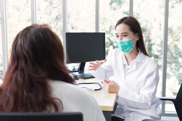 Asiatische Ärztin spricht mit ihrem Patienten, während sie im Krankenhaus eine medizinische Gesichtsmaske trägt