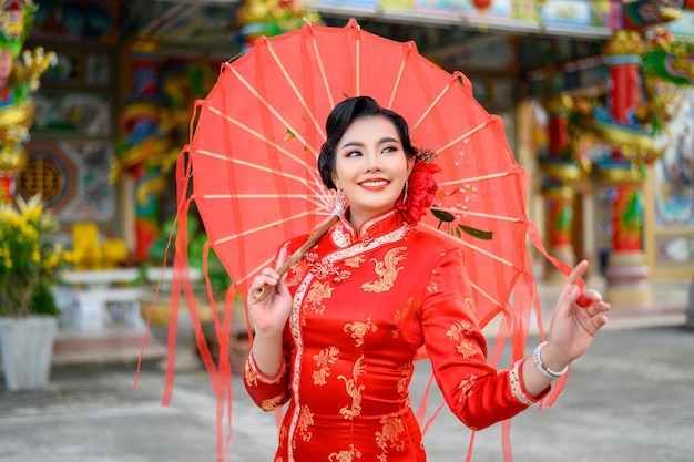 Asiatische schöne Frau des Porträts, die ein Cheongsam trägt, lächelt und posiert mit rotem Regenschirm aus Papier am Schrein am chinesischen Neujahrsfest