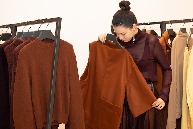 Asiatische schöne Frau im Kleid wählt neue Kollektion auf orangegrünem Erdton-Kleiderständer im Modegeschäft aus, das nur Markennachrichten für den Winterherbst als minimalen Stil öffnet