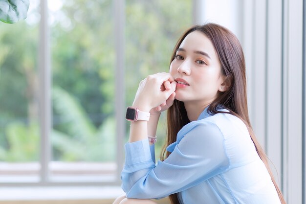 Asiatische schöne Frau trägt blaues Hemd und intelligente Uhr und sitzt in der Nähe des Fensters als Hintergrund.