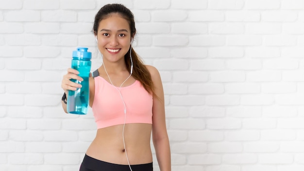 Asiatische schöne Frauen, die Wasserflasche nach dem Spiel Yoga und Übung auf weißem Backsteinmauerhintergrund mit Kopierraum haltenÜbung zum Abnehmen erhöhen die Flexibilität und straffen die Form