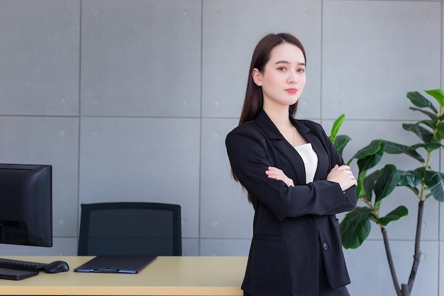 Asiatische schöne Geschäftsfrau im schwarzen Anzug, die glücklich im Büro mit verschränkten Armen steht