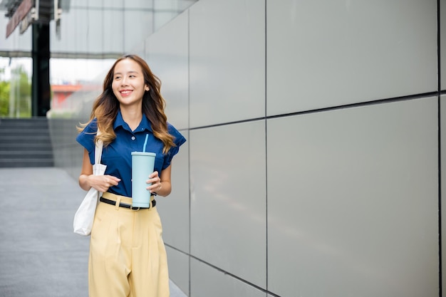 Asiatische schöne Geschäftsfrau, selbstbewusst lächelnd mit Stofftasche, die Stahl-Thermoskanne mit Wasserglas hält, sie geht im Freien auf der Straße in der Nähe des modernen Gebäudebüros spazieren, glückliche Frau, die zur Seite schaut