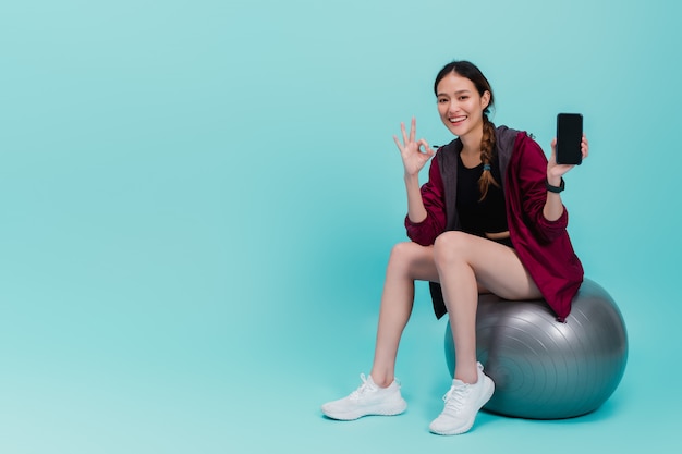 Asiatische schöne glückliche Frau, die Smartphone hält und auf Sitzball nach der Übung lokalisiert auf Blau sitzt