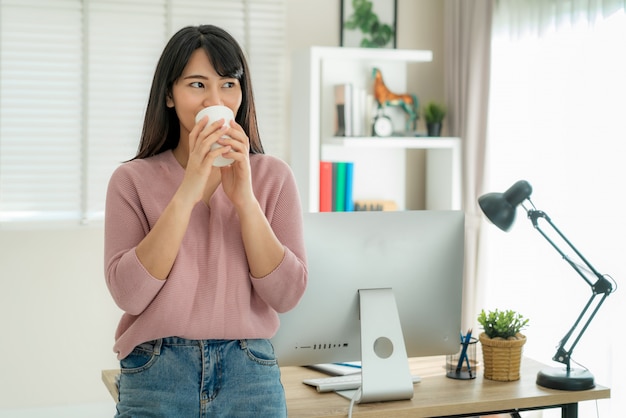 Asiatische schöne junge Frau arbeiten von zu Hause aus am Computer und trinken Kaffee, während sie sich von ihrer Arbeit im Wohnzimmer zu Hause entspannen.