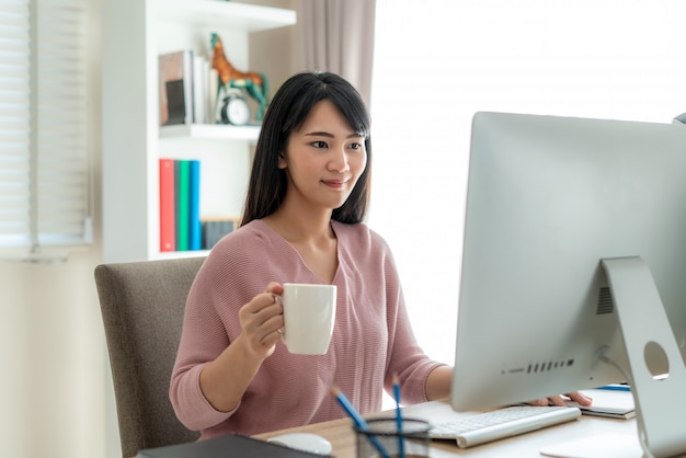 Asiatische schöne junge Frau arbeiten von zu Hause aus am Computer und trinken Kaffee