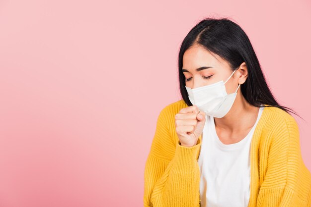 Asiatische schöne junge Frau mit medizinischem Maskenschutz zur Vorbeugung von Infektionen mit Coronavirus, COVID-19 sie hustet niesen, Studioaufnahme isoliert auf rosa Hintergrund, medizinisches Gesundheitskonzept