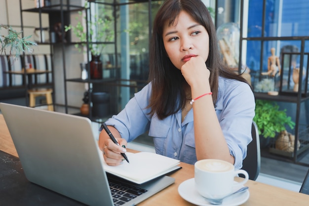 Asiatische Schönheit im blauen Hemd unter Verwendung des Laptops und des trinkenden Kaffees