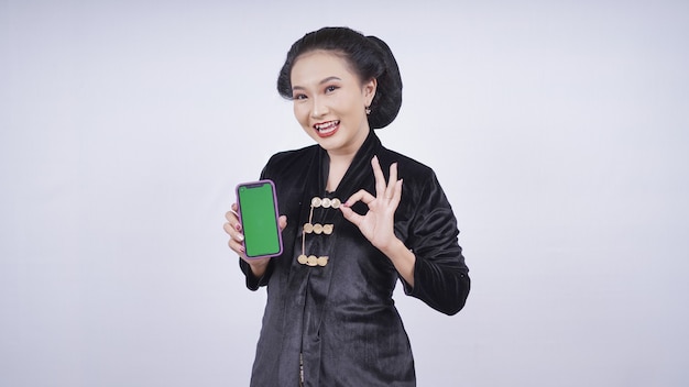 Asiatische Schönheit in Kebaya zeigt Smartphone-Bildschirm-OK-Geste isoliert auf weißem Hintergrund