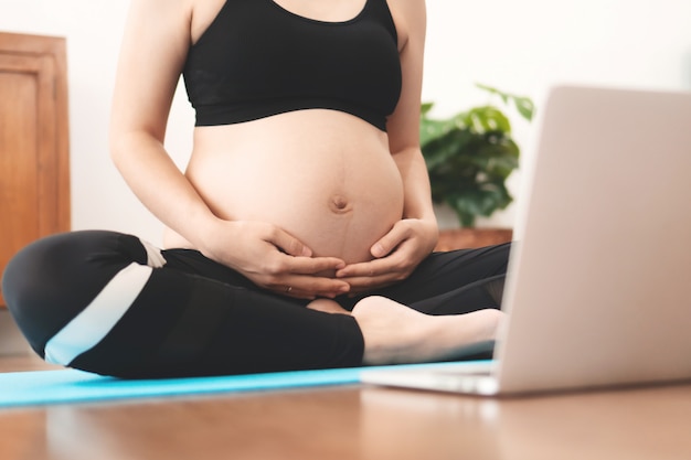 Asiatische schwangere Frau, die Yoga mit Online-Videotraining auf Laptop praktiziert