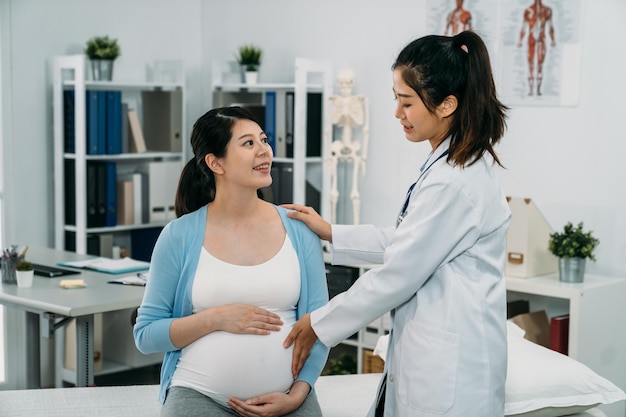 asiatische schwangere Mutter, die auf dem Behandlungsbett sitzt, lächelt die Ärztin an, während sie ihren Bauch berührt und die Gesundheit des Babys im Krankenhaus überprüft.
