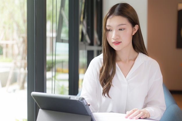 Asiatische selbstbewusste Frauen, die ein weißes Hemd tragen, verwenden ein Tablet zum Arbeiten und es liegen viele Dokumente auf dem Tisch. ihr Gesicht mit dem Lächeln in einem Arbeitszimmer zu Hause.