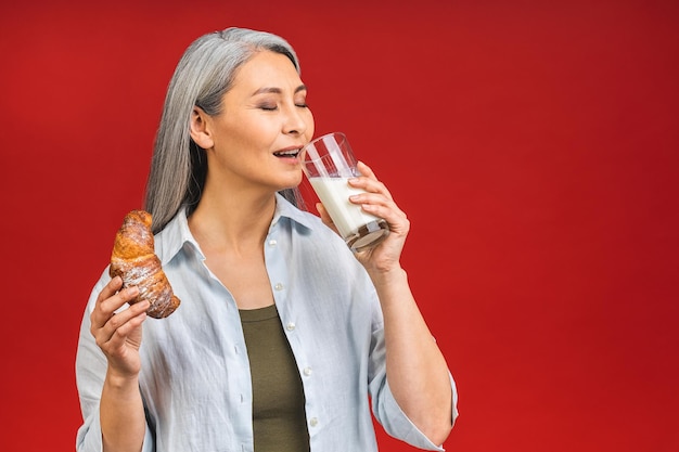 Asiatische Seniorin, reife Frau, die Milch für Kalziumnahrung und ein gesundes Lebensstilkonzept trinkt, das auf rotem Hintergrund isoliert ist Lady isst Croissant