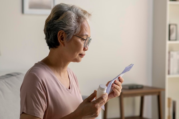 Asiatische Seniorin sitzt auf dem Sofa in ihrem Wohnzimmer und liest das Informationsblatt zu ihrem verschriebenen Medikament