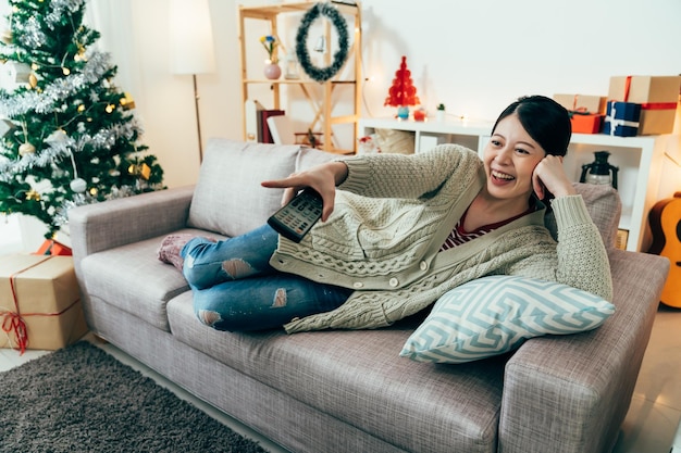 asiatische Stubenhocker zeigt auf den Bildschirm, während sie lustige Fernsehsendungen anschaut. Frau, die auf der Seite auf dem Sofa liegt, lacht herzlich. Indoor-Erholungsideen für die Weihnachtsferien. echte Momente