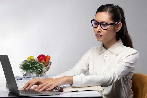 Asiatische Studentin der jungen Universität, die lila Brillenforschung und suchende Informationen auf Computer-Laptop-Notizbuch trägt