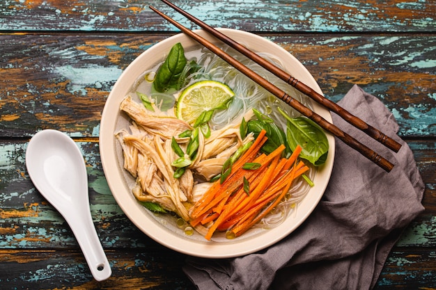 Asiatische Suppe mit Reisnudeln, Hühnchen und Gemüse in Keramikschale, serviert mit Löffel und Stäbchen auf rustikalem Holzhintergrund von oben, chinesische oder thailändische Küche