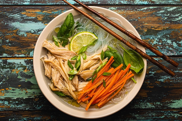 Asiatische Suppe mit Reisnudeln, Hühnchen und Gemüse in Keramikschale, serviert mit Löffel und Stäbchen auf rustikalem Holzhintergrund von oben, chinesische oder thailändische Küche