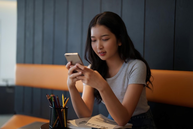 Asiatische Teenager-Studentin, die Smartphone benutzt, um mit Freunden zu chatten und in sozialen Medien zu surfen, nach dem Studium und dem Lesen von Informationen in Papierarbeit, um über Bildung und Wissen in einem Café zu lernen