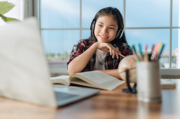 Asiatische Teenager-Studentin trägt drahtlose Kopfhörer online mit Zoom-Meeting, glückliche junge Frau lernt Sprache hören Vorlesung Webinar ansehen Notizen schreiben Blick auf Laptop-Fernbildung.