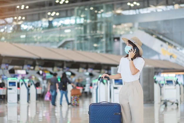 Asiatische Touristin, die Gesichtsmaske trägt und ein mobiles Smartphone im Flughafenschutz verwendet Coronavirus-Krankheitsinfektion Reisezeit nach dem Konzept der Impfstoff-Booster-Dosis