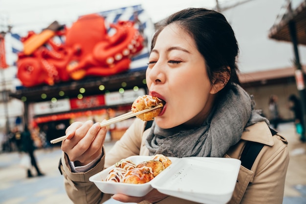 asiatische touristin, die heiße takoyaki mit essstäbchen probiert. junge elegante dame, die vor dem straßenhändlergeschäft steht, das tacoyaki verkauft. Mädchen, das köstliche Oktopusbällchen isst.