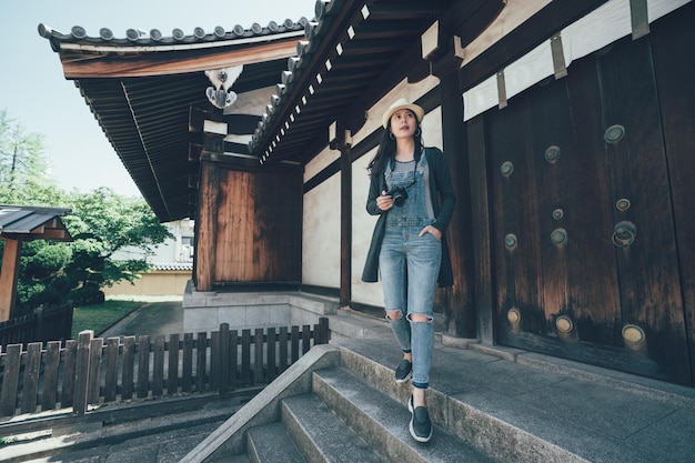 asiatische touristin in voller länge geht von der treppe mit kamera außerhalb des shitennoji-tempels osaka japan hinunter. Junges Mädchen, das mit den Händen in der Tasche spazieren geht und ein Foto macht, besuchen Sie den Shinto-Look.