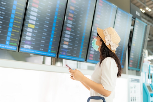 Asiatische Touristin mit Gesichtsmaske, die den Flug vom Ankunftsabflugbrett im Flughafenterminal überprüft