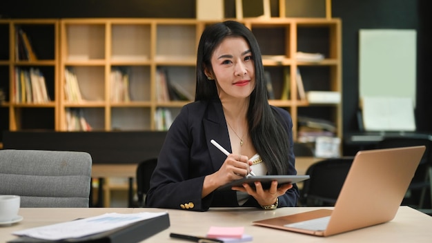 Asiatische Unternehmerin, die in ihrem Büro vor einem Laptop sitzt und ein digitales Tablet verwendet