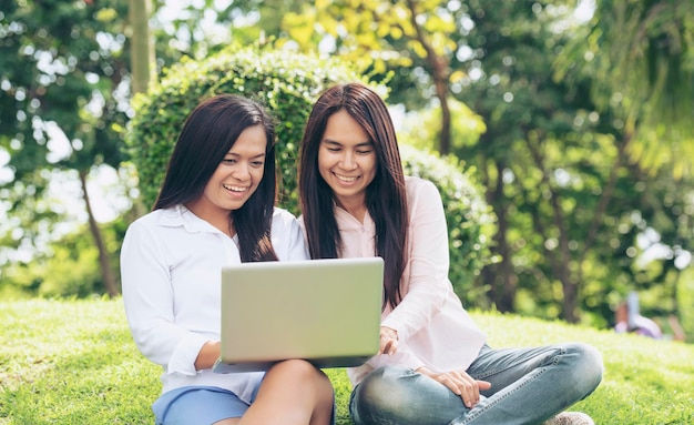 Asiatische zwei Frauen sprechen mit Laptop sitzt auf der grünen Wiese im Park Freundschaft Mädchen chatten und sprechen Blick auf Laptop-Planung tun kleine Unternehmen grünen Garten Junge schöne Frauen lachen miteinander reden