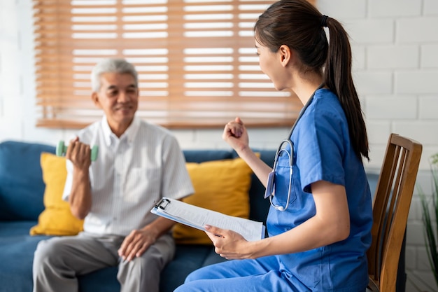 Asiatischer älterer älterer Mann macht Physiotherapeut mit Unterstützung von Krankenschwester Reifer älterer männlicher Patient sitzt auf dem Sofa im Wohnzimmer mit Hanteln Workout Übung mit Betreuer zu Hause Pflege