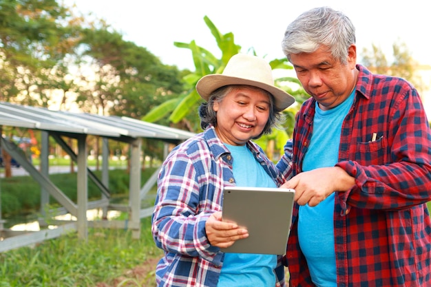 Asiatischer älterer Bauer hält ein Tablet in der Hand, mit dem das Solarpanel gesteuert wird, um Strom zu verbrauchen