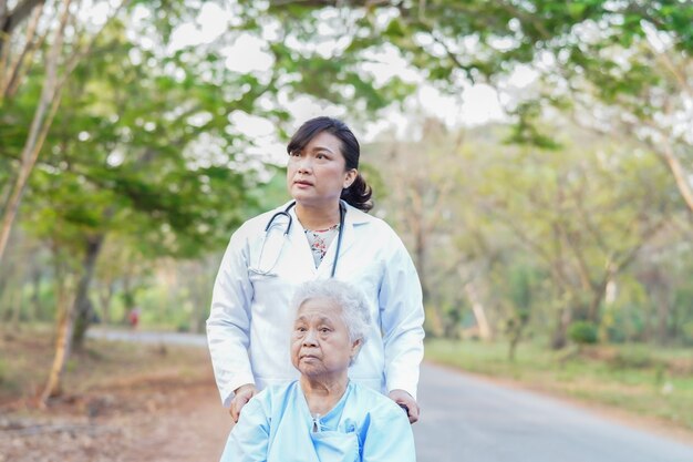 Asiatischer älterer Frauenpatient der Doktorhilfe und -sorgfalt, der auf Rollstuhl am Park sitzt.