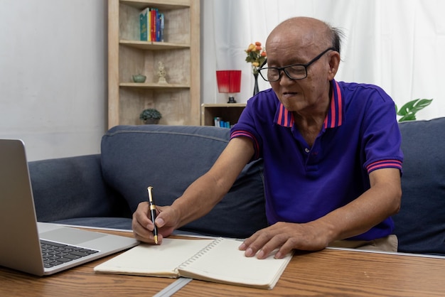 Asiatischer älterer Mann, der einen Stift hält, schreibt eine Nachricht und einen Computer-Laptop auf dem Tisch am Sofa