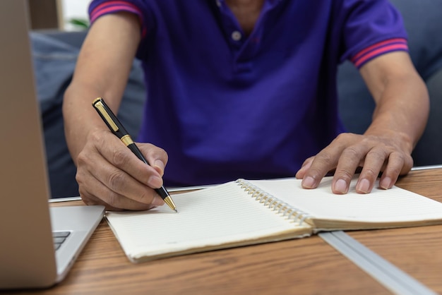 Asiatischer älterer Mann, der einen Stift hält, schreibt eine Nachricht und einen Computer-Laptop auf dem Tisch am Sofa