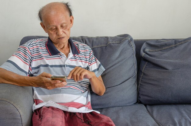 Asiatischer älterer Mann, der sein Telefon auf dem blauen Sofa im Haus betrachtet