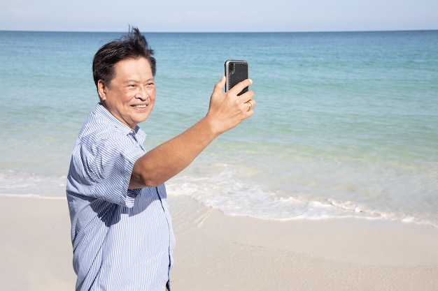 Asiatischer älterer Mann nimmt Selfie mit Handy am Strand