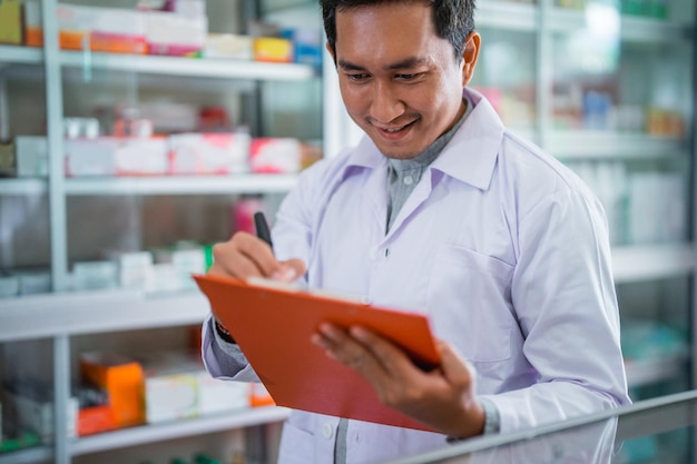 Asiatischer Apotheker in Uniform, der während der Registrierung in der Apotheke mit der Schreibtafel arbeitet