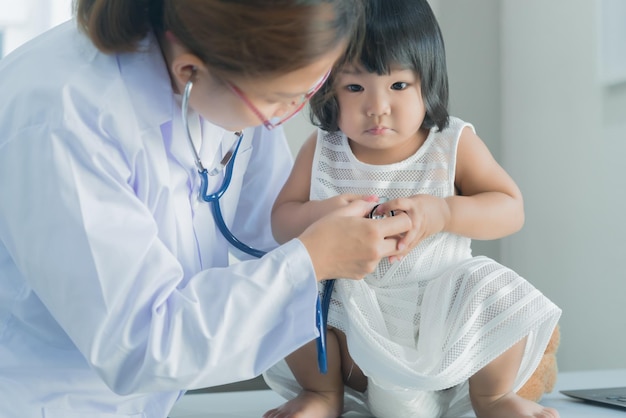 Asiatischer Arzt, der ein Stethoskop verwendet, um seine Atmung und das Herz eines hübschen Mädchens zu überprüfen, überprüft die Gesundheit von Kindern in Thailand