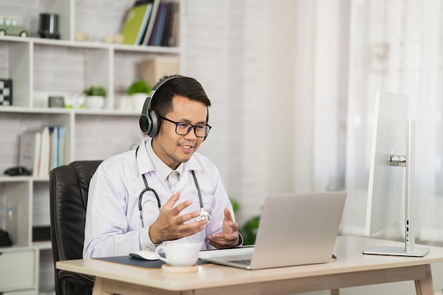 Asiatischer Arzt, der Kopfhörer trägt und einen Laptop verwendet, um den Patienten per Videoanruf im Home Office zu beraten. Telemedizin- und Gesundheitskonzept